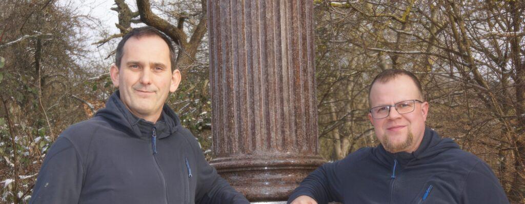 Zwei Männer stehen lächelnd neben einer Säule in der Natur.